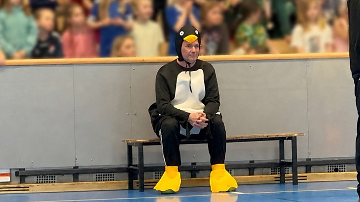 Lokalt i:s pingvinklädde krönikör bänkade sig själv i handbollsdebuten i Maserhallen. (Obs: Bilden har redigerats via ett AI-verktyg, som gjort barnen på läktaren oskarpa). Foto: Ingela Sönnert